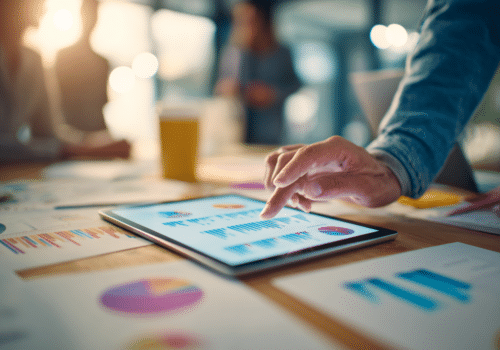 Une personne consulte des graphiques et tableaux de performance marketing sur une tablette posée sur une table de réunion, entourée de documents imprimés présentant des données et statistiques. Scène en environnement professionnel, centrée sur l’analyse de données
